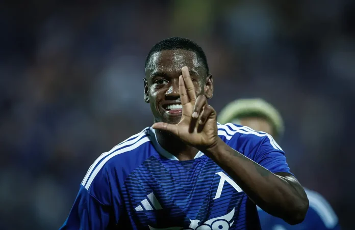 Soccer player in a blue jersey celebrating on the field, holding up three fingers toward the camera.