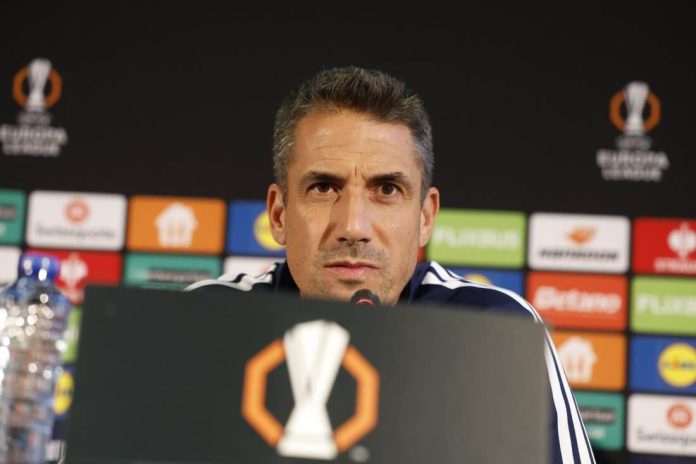 Sports coach at a press conference speaking into a microphone, backdrop with sponsor boards and a trophy logo on the podium