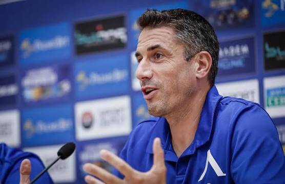 Man in a blue polo shirt speaking at a press conference and gesturing with his hands.