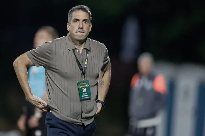 Man in a striped polo with a lanyard and badge, standing with hands on hips during an event, surprised expression.
