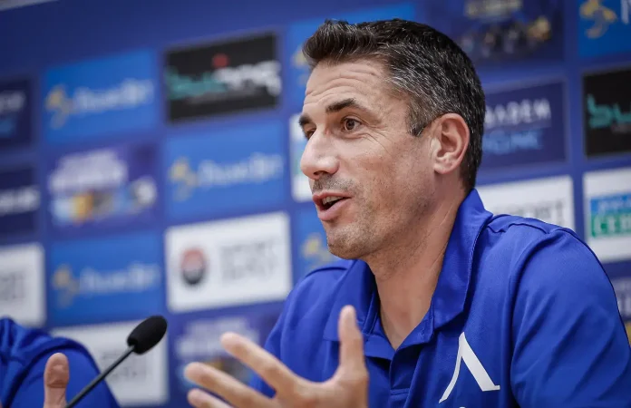 Man in a blue polo speaking at a press conference, gesturing with his hands in front of a backdrop of logos and a microphone nearby.