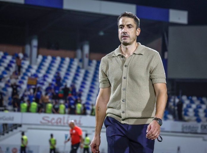 Man in a beige short-sleeve polo and navy pants walks across a stadium field with blurred spectators in the stands behind him.