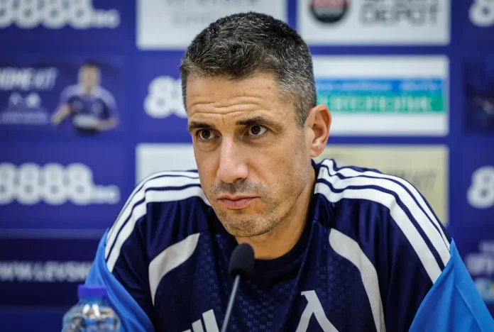 Man in a navy sports tracksuit at a press conference, speaking into a microphone against a blue backdrop.
