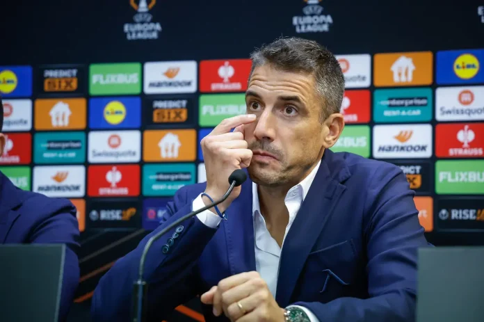 Man in a blue suit at a press conference, touching his eye while speaking into a microphone, backdrop of sponsor logos.