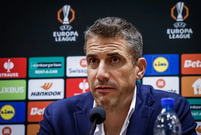Man in a navy suit at a press conference with a Europa League backdrop and sponsor logos behind him, speaking into a microphone.