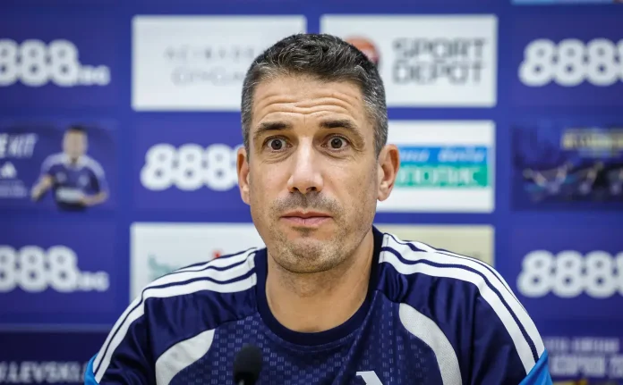Man in a blue sports jersey at a press conference, facing the camera with microphones in front of a blue sponsor backdrop.