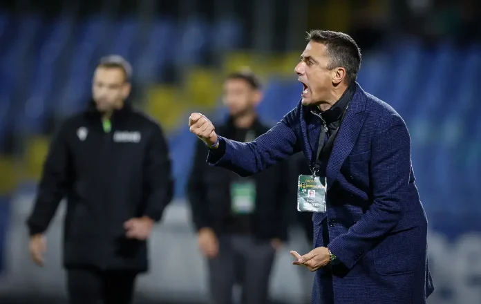 Coach in a navy blazer shouting and gesturing on the sideline during a game, badge hanging from his neck.