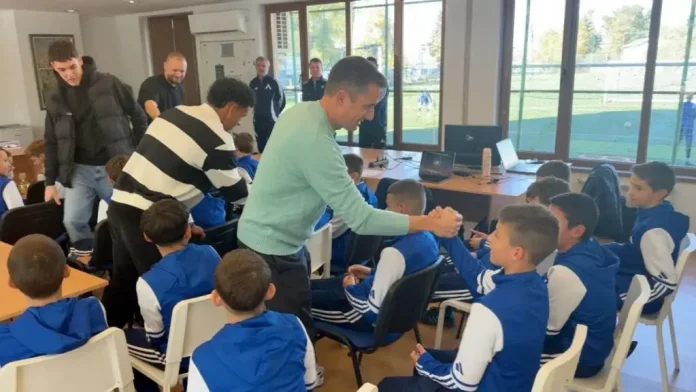 A man in a mint sweater shakes hands with a seated student while others stand nearby in a classroom filled with blue-uniformed kids and laptops on desks.