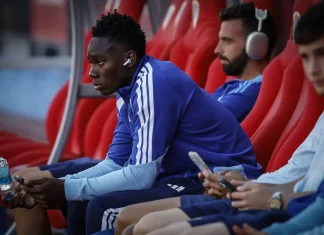 Камерунски национал отново в плановете на Хулио Веласкес Soccer players sit on a bench in blue training gear, one wearing AirPods and holding a water bottle and phone.