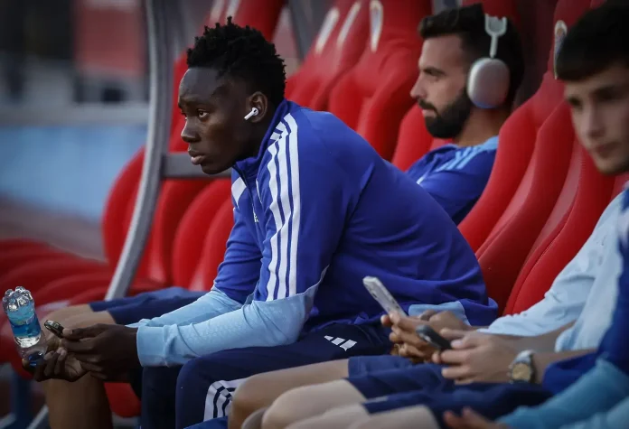 Soccer players sit on a bench in blue training gear, one wearing AirPods and holding a water bottle and phone.