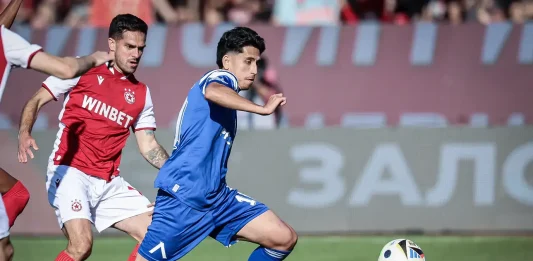 Кой ще замени Асен Митков срещу Черно море? Blue-shirted player runs toward the ball while a red-clad defender pursues on a grass soccer field.