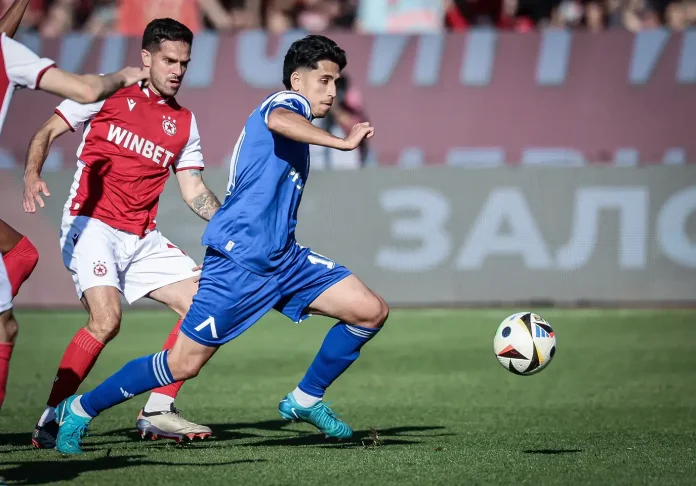 Blue-shirted player runs toward the ball while a red-clad defender pursues on a grass soccer field.