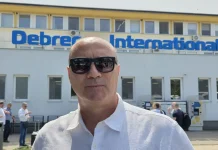 Кокала: Левски пак ще е шампион Man in a white shirt wearing dark sunglasses stands in front of a building with a blue 'Debrecen International' sign above the windows.