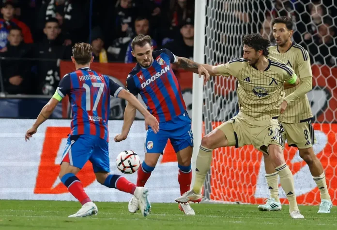 Soccer players in blue-red striped kits contest the ball near the goal as a goalkeeper in light gold stands ready behind them