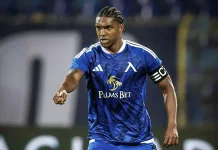 Кристиан Макун: биография, кариера и статистика Soccer player in blue kit with 'Palms Bet' sponsor, wearing captain's armband, pointing forward on the field.
