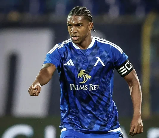 Кристиан Макун: биография, кариера и статистика Soccer player in blue kit with 'Palms Bet' sponsor, wearing captain's armband, pointing forward on the field.