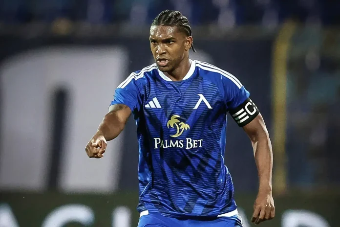 Soccer player in blue kit with 'Palms Bet' sponsor, wearing captain's armband, pointing forward on the field.