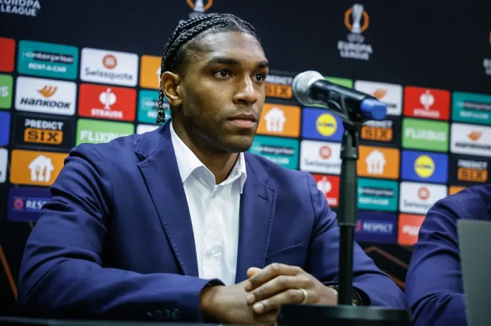Man in a navy suit at a press conference, seated behind a microphone with sponsor boards in the background
