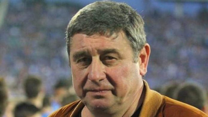 Close-up of a middle-aged man with short gray hair wearing an orange jacket at a crowded event.