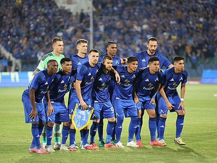 A soccer team in blue uniforms posing for a group photo on the field with a crowd in the stands behind them.