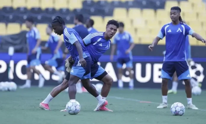 Soccer players in blue training kits dribbling balls on a green field during practice with yellow stadium seats in the background