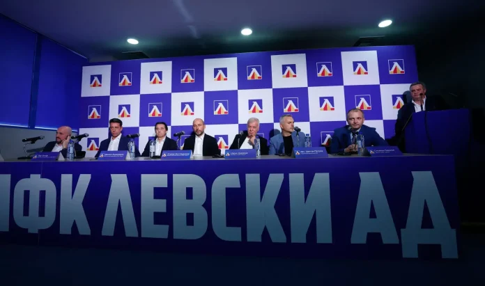 Panel of seven men seated at a press conference table with microphones in front of a blue checkered backdrop featuring a logo and large Cyrillic lettering in the foreground.