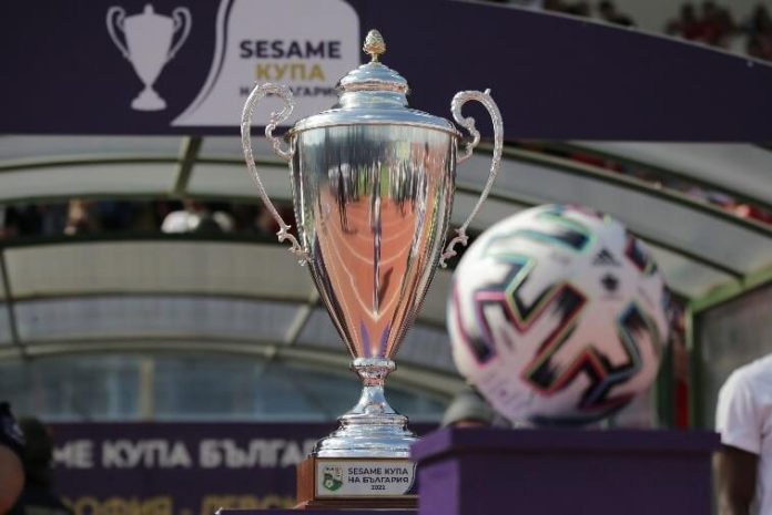 Shiny silver trophy with ornate handles on a wooden base, in a stadium setting, with a colorful soccer ball nearby and 'Sesame Cup' visible on the base.