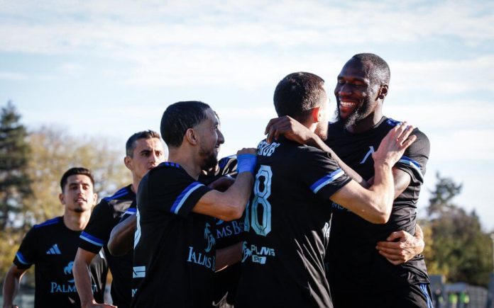 Soccer teammates in black uniforms hug and celebrate on a sunny field.