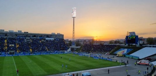 Левски има специален спомен от 15 октомври Football stadium at sunset with players on the green pitch and a large crowd in blue stands.