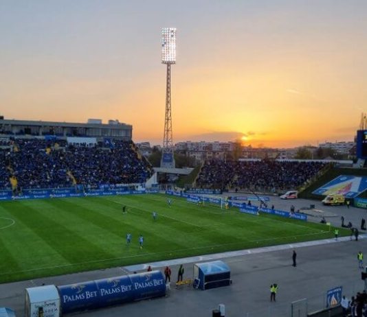 Левски има специален спомен от 15 октомври Football stadium at sunset with players on the green pitch and a large crowd in blue stands.
