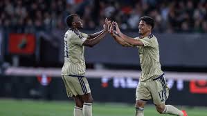Two soccer players in beige kits high-five each other on the field, celebrating with a cheering crowd in the background.
