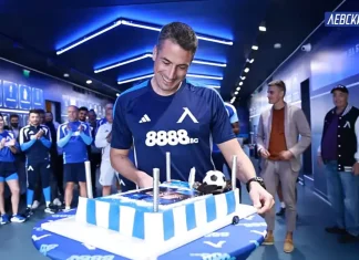 Левски изненада Веласкес за рождения му ден Man in a blue team jersey cuts a large blue-and-white striped cake while teammates applaud in a hallway.