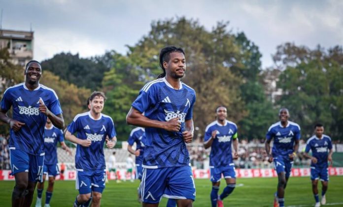 Soccer players in blue uniforms jogging on a grassy field during a match, with trees and a stadium in the background.
