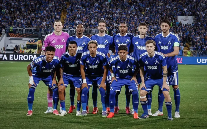 A soccer team in blue kits poses for a group photo on the pitch before a match, with players kneeling in front and standing behind them as a cheering crowd watches.