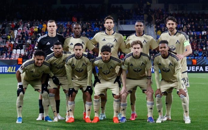 Soccer team in beige away kits posing for a group photo on the field before a match, with stands in the background.