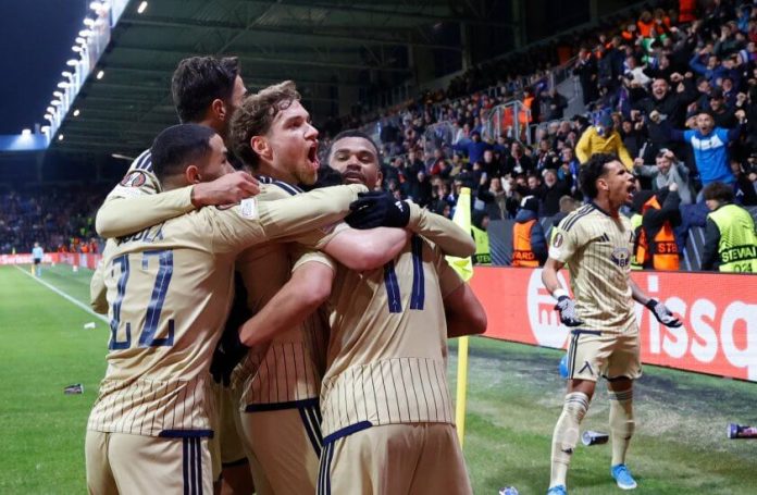 Soccer players in beige uniforms hug in celebration on the pitch as fans cheer in the stands behind them.
