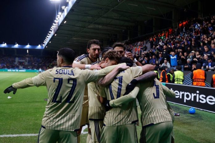 Soccer players in beige uniforms huddle together in celebration on the pitch as spectators cheer in the stands in the background.