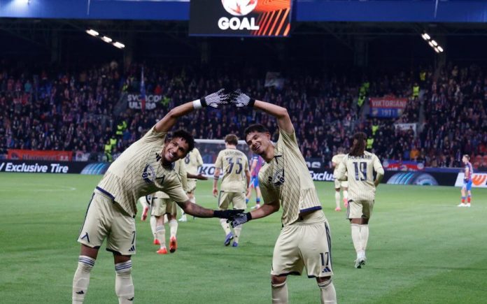 Two soccer players in cream jerseys form a heart with their arms, celebrating a goal on a crowded stadium field.