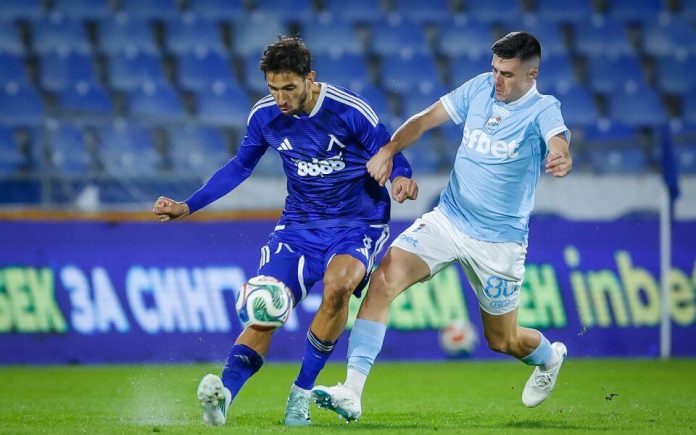 Two soccer players in blue uniforms fight for the ball on a grassy field during a match as a light-blue kit defender challenges a dark-blue attacker.