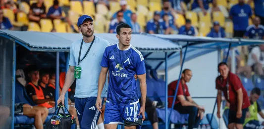 Левски няма да бърза с Бурас Soccer player in blue kit (number 47) stands by the dugout with a staff member carrying a medical bag, stadium seating in yellow behind.
