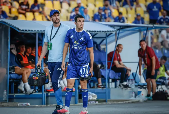 Soccer player in blue kit (number 47) stands by the dugout with a staff member carrying a medical bag, stadium seating in yellow behind.