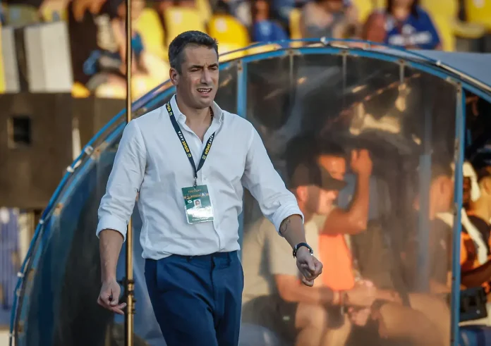 Coach walking out of team dugout at a stadium, wearing a white shirt, blue pants, and a credential badge on a lanyard.