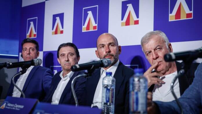 Four men in suits sit at a panel with microphones, water bottles, and a blue backdrop featuring logos behind them (press conference).