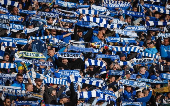 Crowd of football fans primarily in blue and white, holding scarves and cheering in a stadium stands.