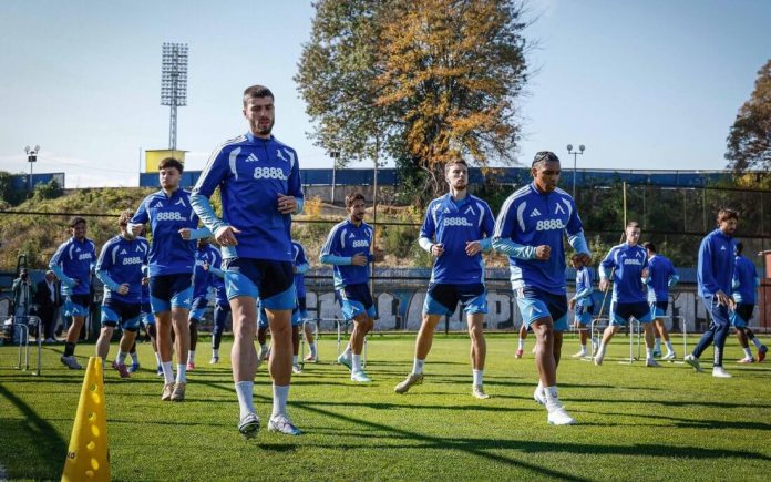 Soccer players in blue uniforms run through a training drill on a sunny green field, with cones and hurdles in front and stadium lights in the background.