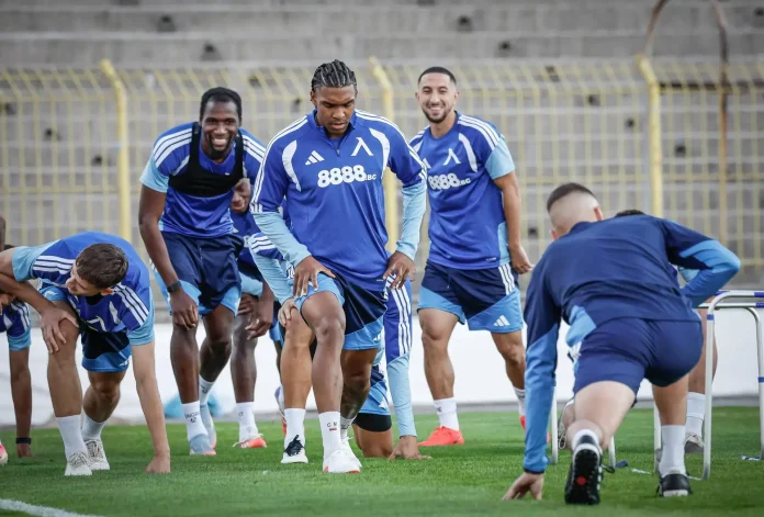 Soccer players in blue uniforms sprinting from the starting line during a drill on a green field, stadium seats in the background.