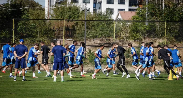 Soccer players in blue uniforms jog across a grassy field during practice, with a chain-link fence and urban buildings in the background.