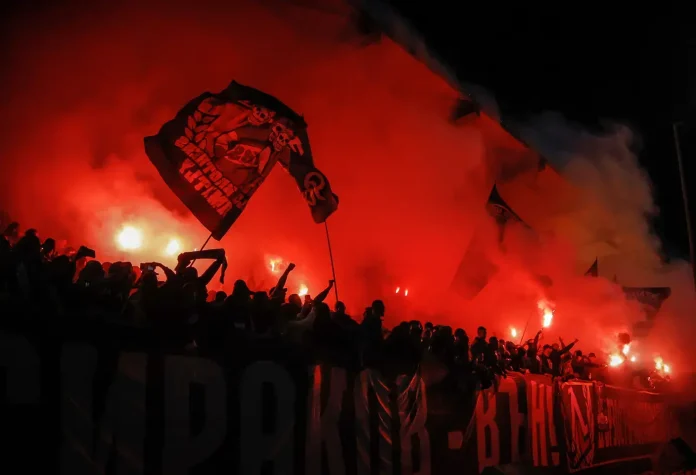 Crowd of football fans in a stadium, waving banners and lighting red flares with thick red smoke filling the scene.
