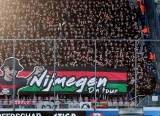 Левски получава неочакван бонус от привържениците на НЕК Crowd of football fans packed in the stands behind a metal barrier, with a large banner featuring a cartoon man and bold lettering.