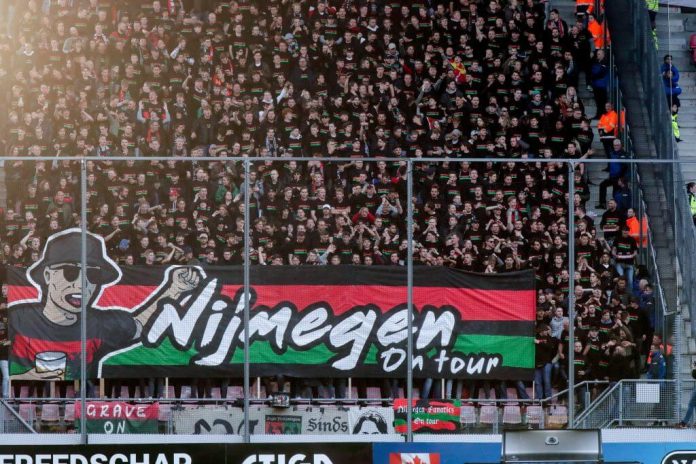 Crowd of football fans packed in the stands behind a metal barrier, with a large banner featuring a cartoon man and bold lettering.